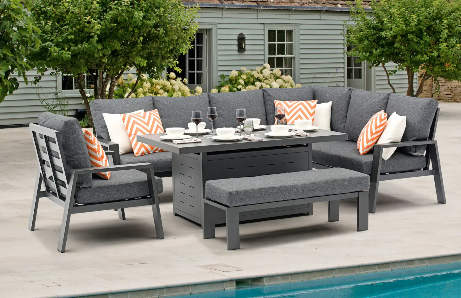 Modern grey Bramblecrest L-shaped garden sofa set with orange cushions, table, fire pit on patio by pool.