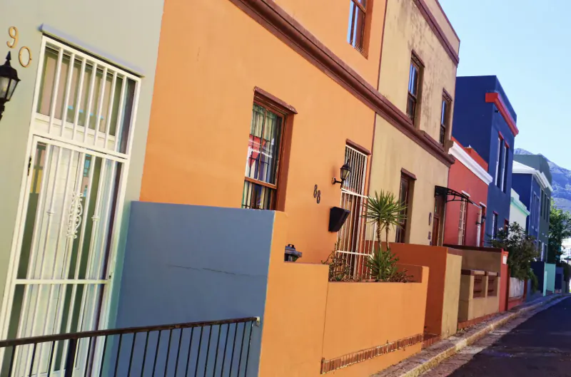 Colorful Cape Malay houses in vibrant hues of orange, teal, and red line a narrow street with mountain backdrop.