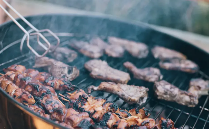 Grilled meat skewers and chops on smoky outdoor barbecue grill with tongs.