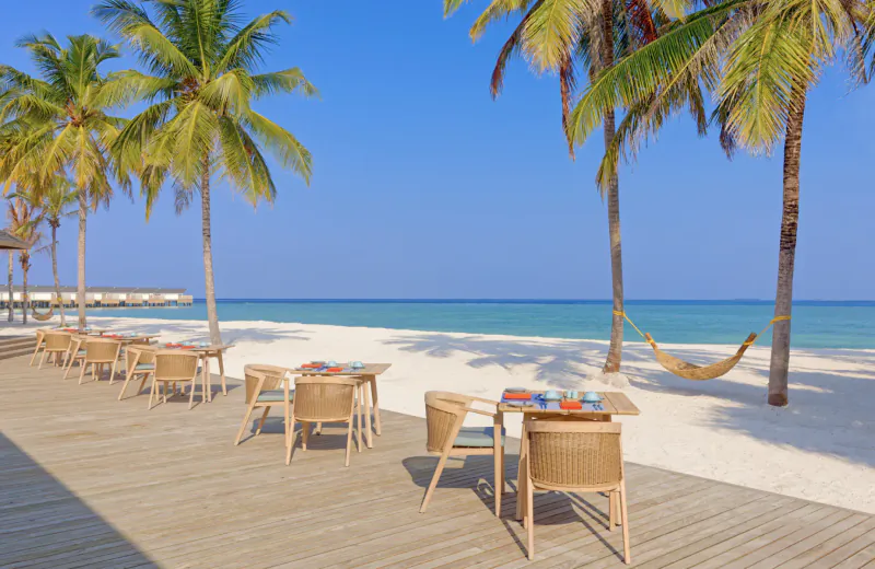 Wooden deck with wicker chairs and tables at Amari Raaya Maldives beach resort, palm trees and turquoise ocean beyond