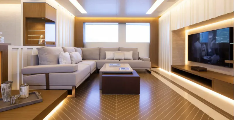 Luxurious yacht lounge with L-shaped gray sofa, brown coffee table, wall-mounted TV, wood floors, and ambient lighting.