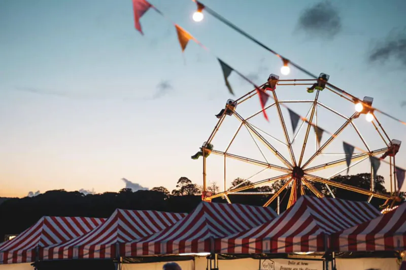 Lit Ferris wheel and red-striped tents with colorful flags at Travelling Feast Festival during twilight.