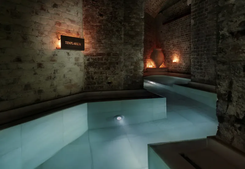 Aire Spa: dimly lit vaulted brick room with turquoise hot tub, 'Aire' sign, urns, and ambient lights.