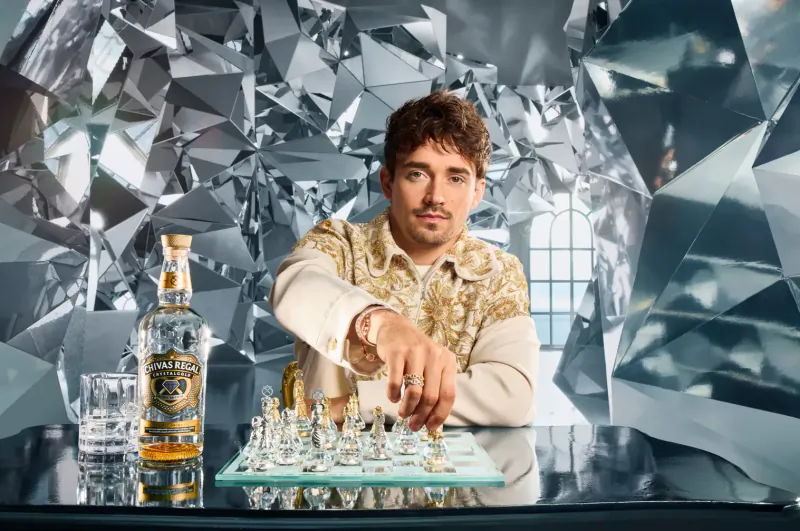 Man in gold shirt thoughtfully moves chess piece on glass board with Luxuria tequila bottle beside him, against shiny silver backdrop.