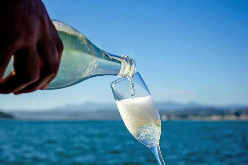 Hand pouring sparkling wine from green bottle into flute glass on ocean sailing charter with sea and mountains backdrop.