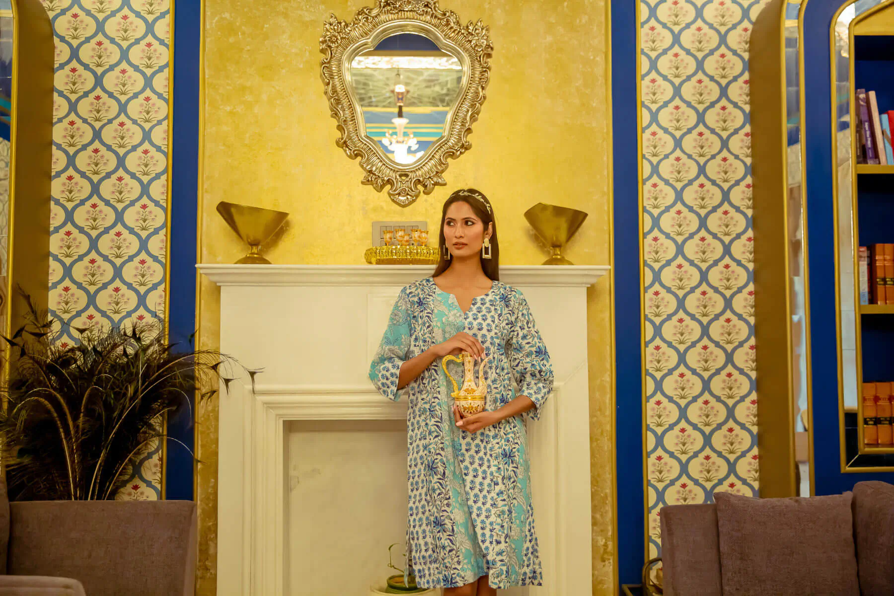 Woman in blue printed natural fabric dress holding cup, standing by ornate gold fireplace in luxurious room with blue wallpaper.