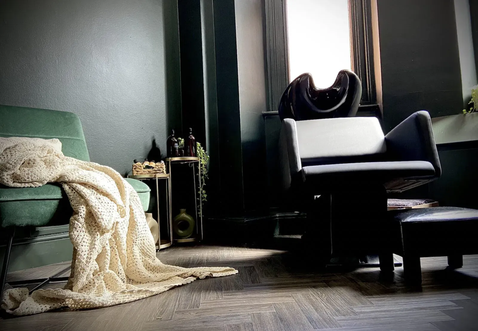 Cozy Jivan Room interior: green velvet sofa with white knit throw, black leather chair, ottoman, plants, and window light.