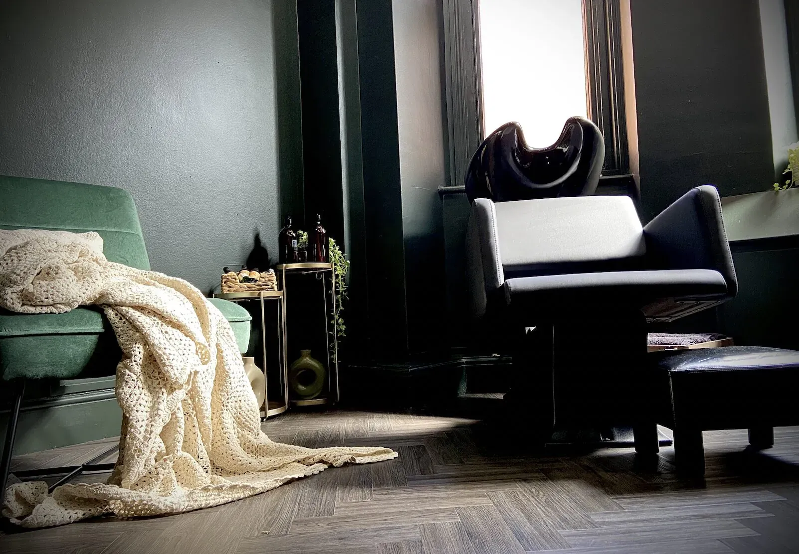 Cozy Jivan Room interior: green velvet sofa with white knit throw, black leather chair, ottoman, plants, and window light.
