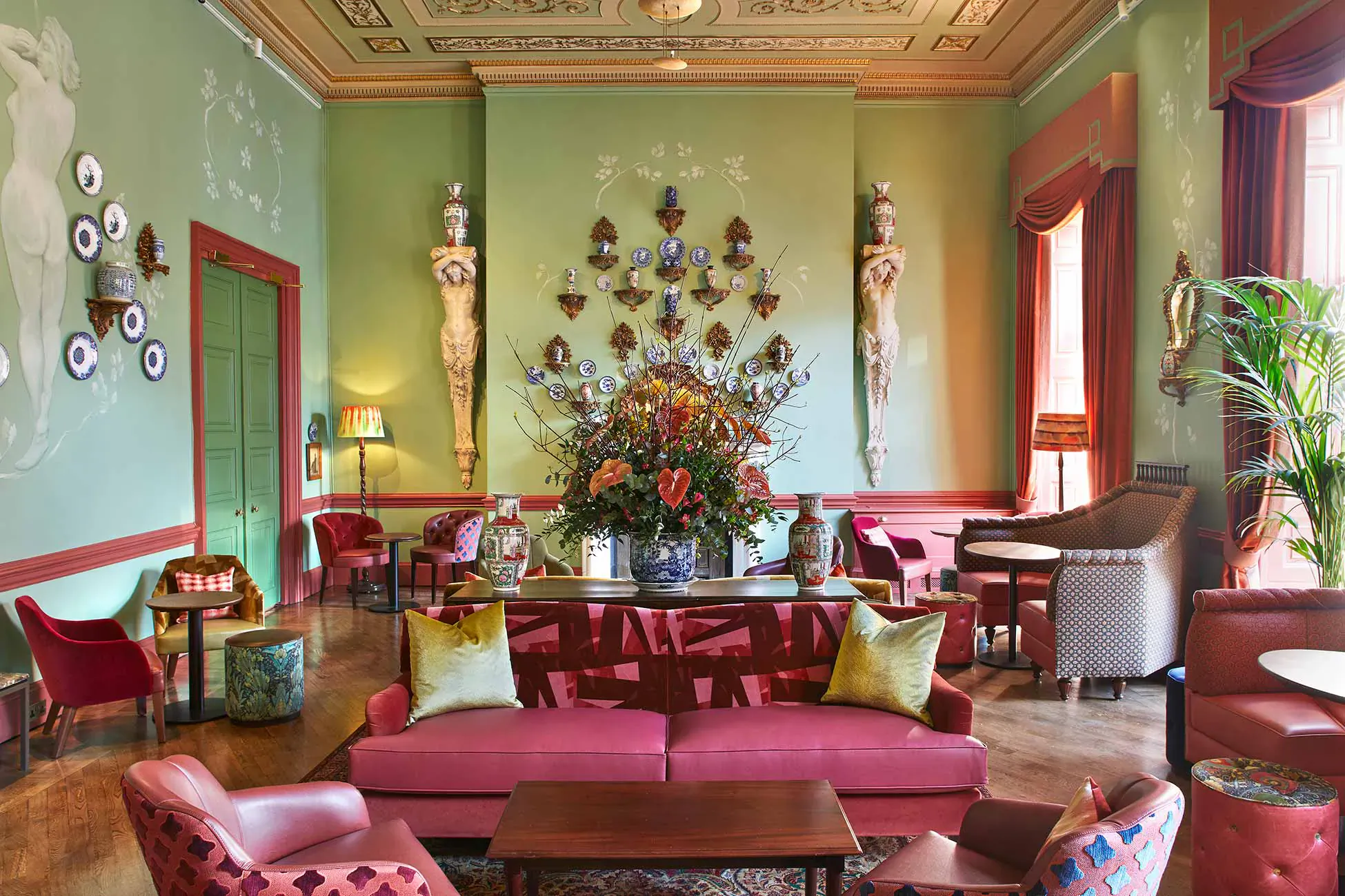 Elegant lounge in Home House club with green walls, ornate statues, large floral centerpiece, and plush red chairs.