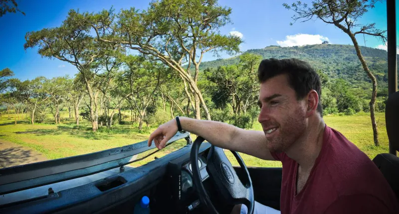 Man with short dark hair smiling and pointing from driver's seat of jeep in African savanna with acacia trees and mountains