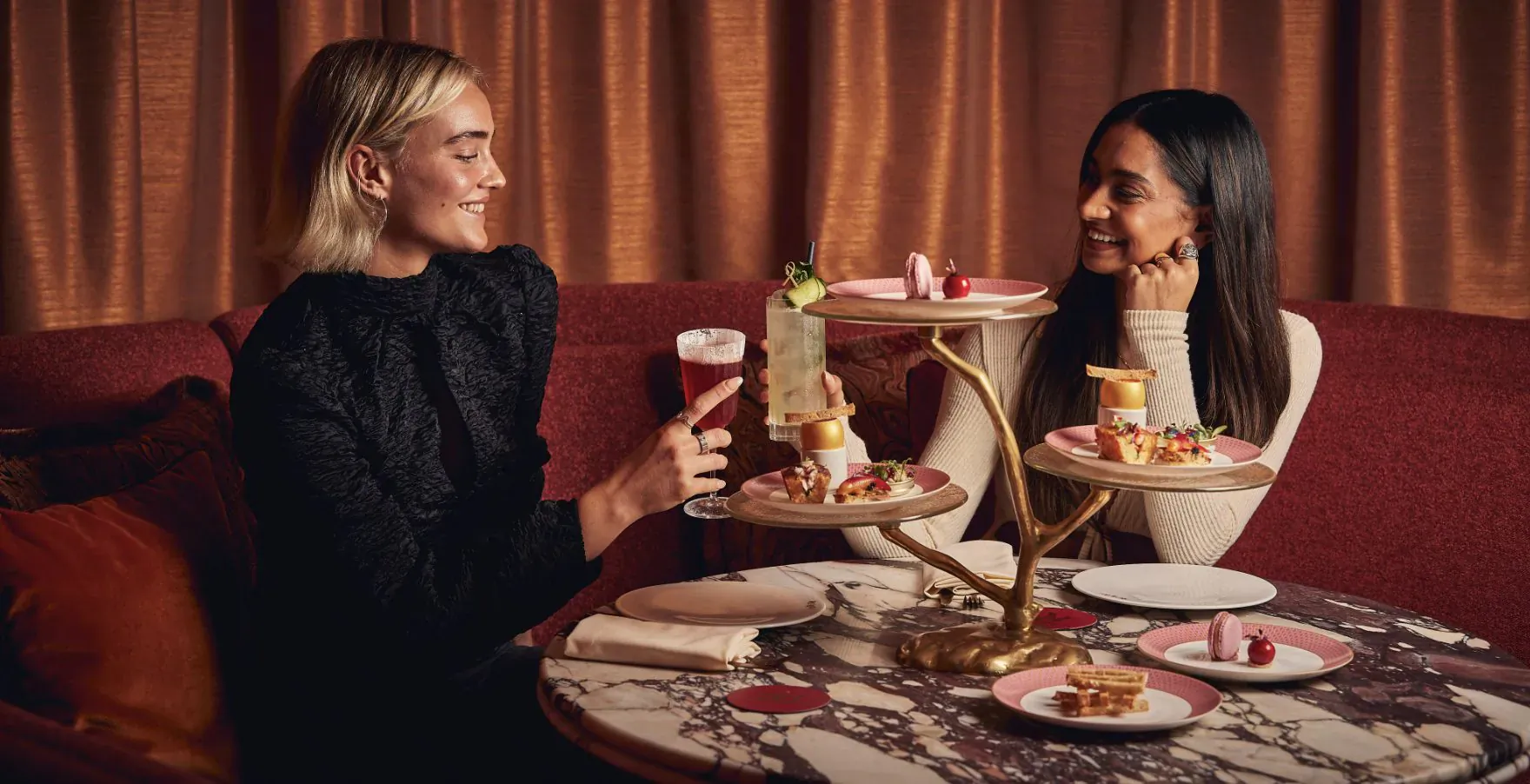 Two women smiling and enjoying afternoon tea at Revery Bar, with tiered stand of pastries, cocktails, and garnishes on marble table.