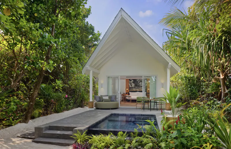 Amari Raaya Maldives white peaked-roof overwater villa with private infinity pool, open doors to bedroom, surrounded by lush tropical greenery under blue sky.