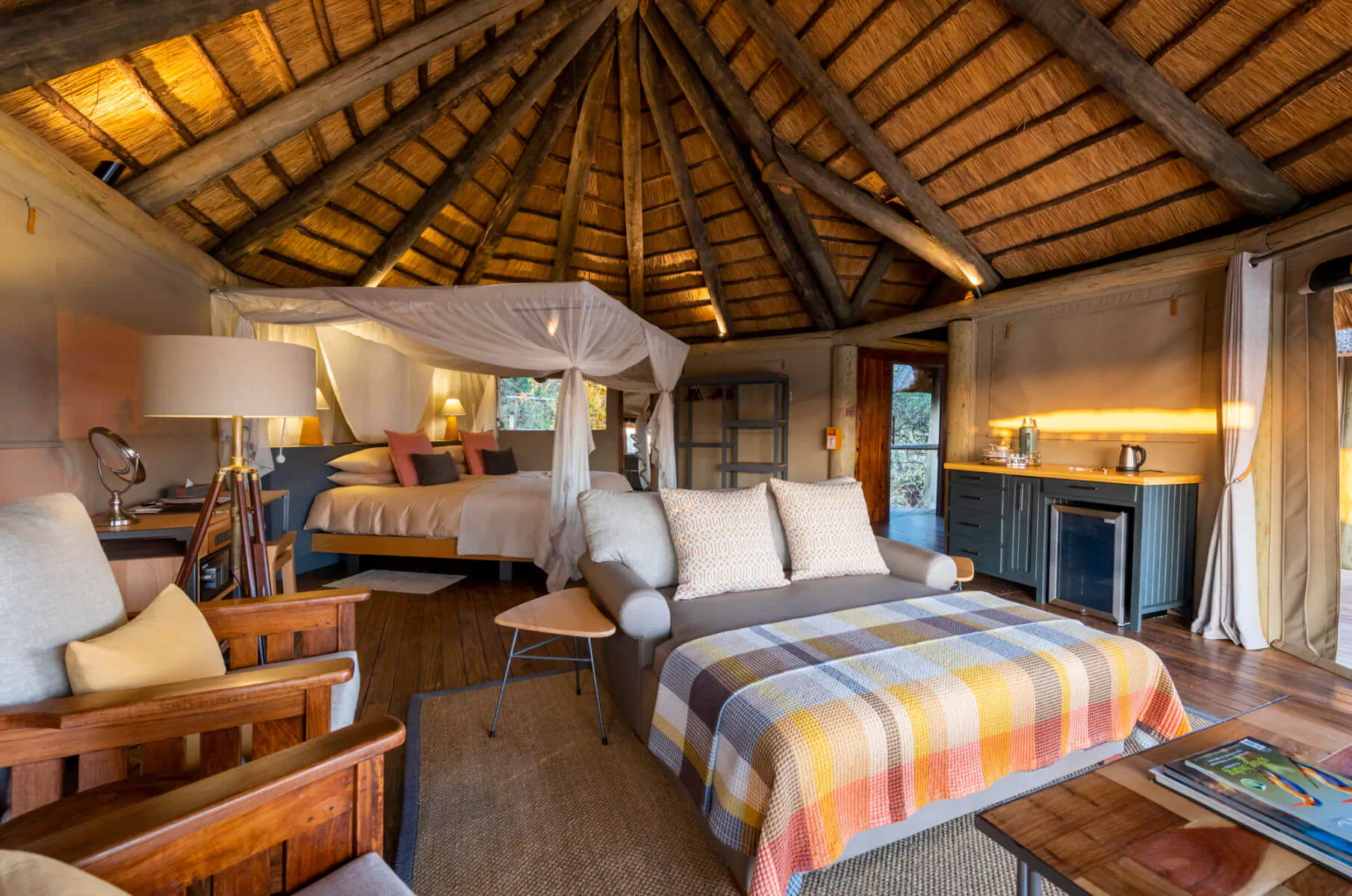 Luxurious rondavel bedroom at Lion Camp with thatched ceiling, mosquito-net canopy bed, wooden chairs, and warm lighting.