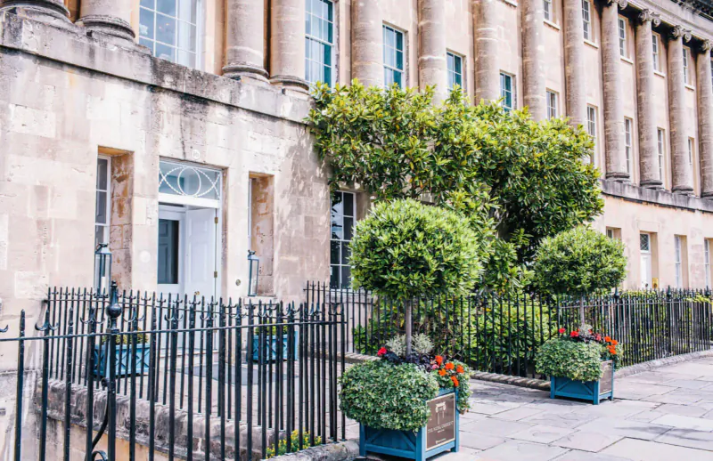 The Royal Crescent in Bath: elegant Georgian facade with columns, climbing vines, topiary shrubs, and flower planters along iron fence.