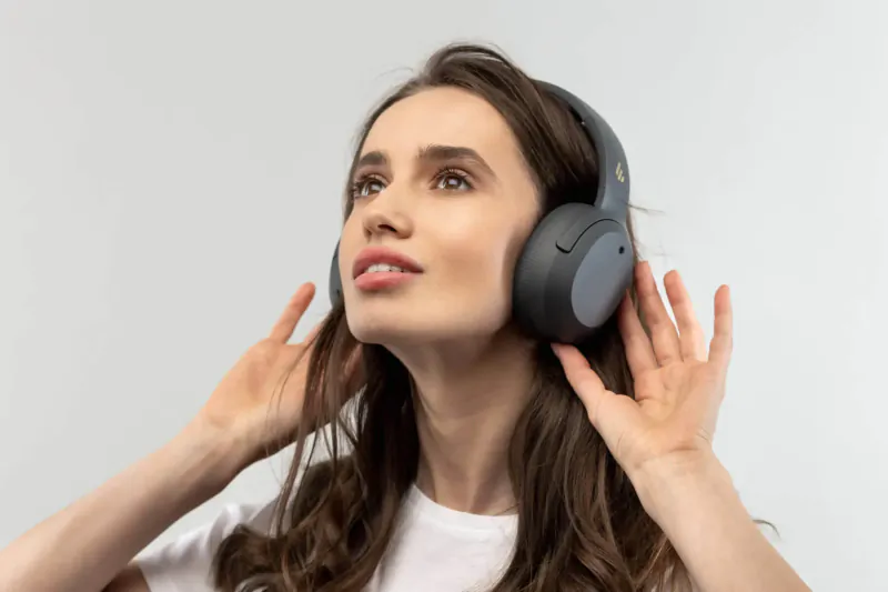Woman with long brown hair wearing gray Sony WH-1000XM4 headphones, hands raised, looking up on white background