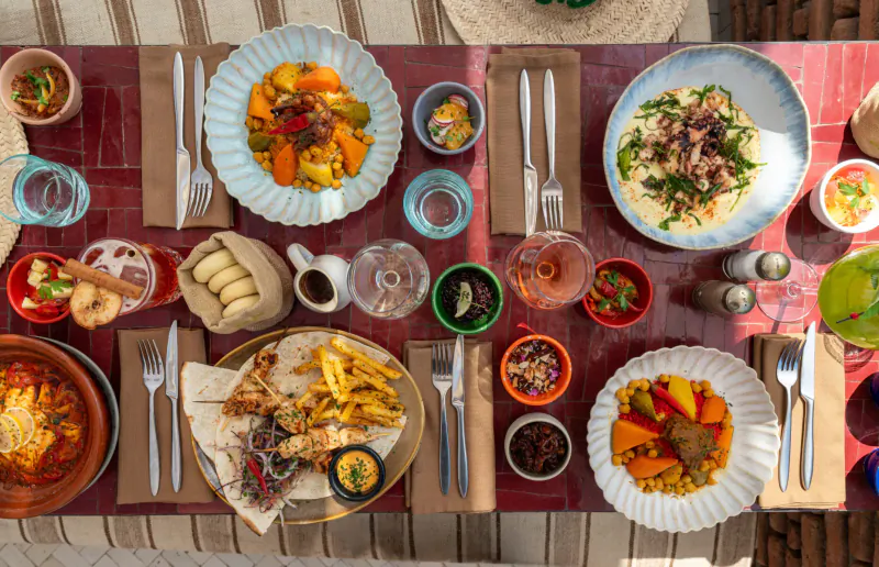 Overhead view of vibrant Moroccan rooftop feast: tagine, couscous, veggies, fries on red table.