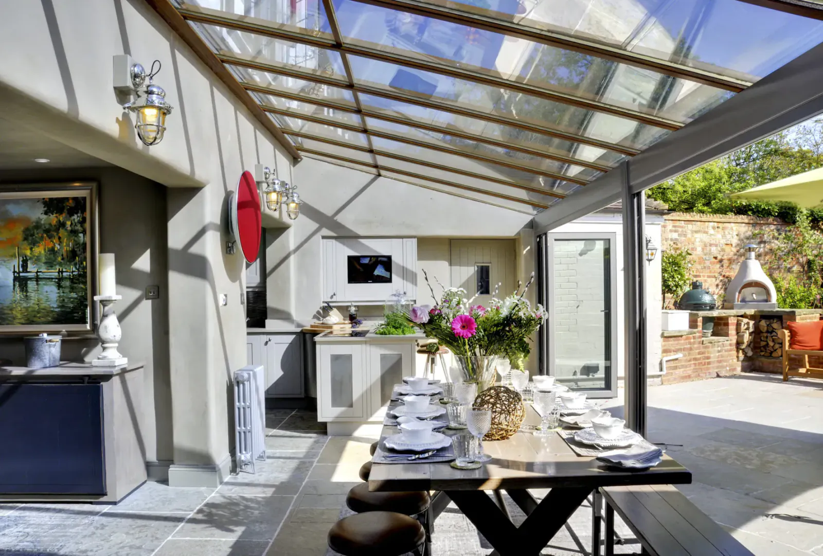 Bright conservatory kitchen-diner in luxury cottage with skylights, set table, flowers, and outdoor BBQ patio.