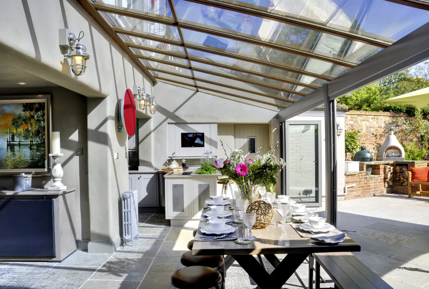 Bright conservatory kitchen-diner in luxury cottage with skylights, set table, flowers, and outdoor BBQ patio.