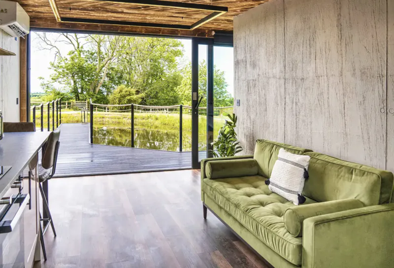 Modern luxury glamping pod interior with green velvet sofa, kitchen bar, and open doors to wooden deck over pond amid green fields.