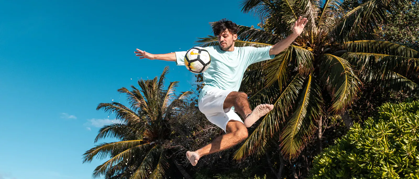 Young man in light blue shirt and shorts leaps kicking yellow soccer ball amid palm trees at beach resort