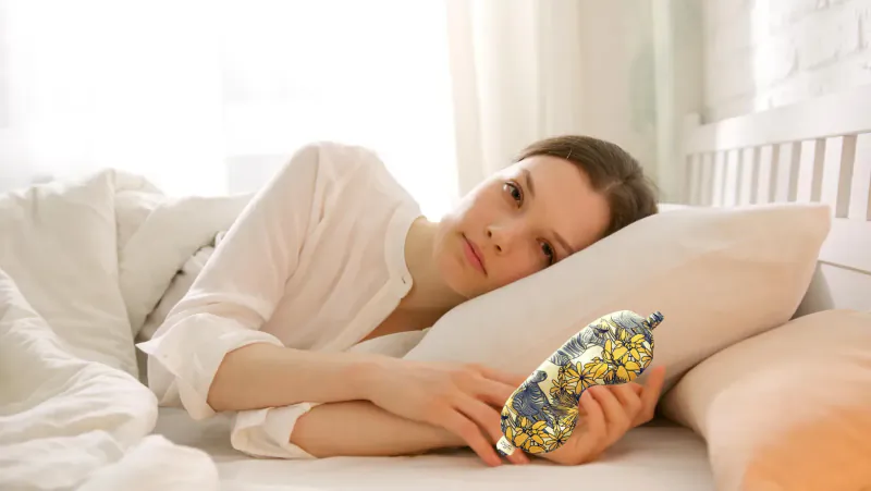 Woman lying in bed, tired expression, holding yellow banana-shaped pillow, soft bedroom light.