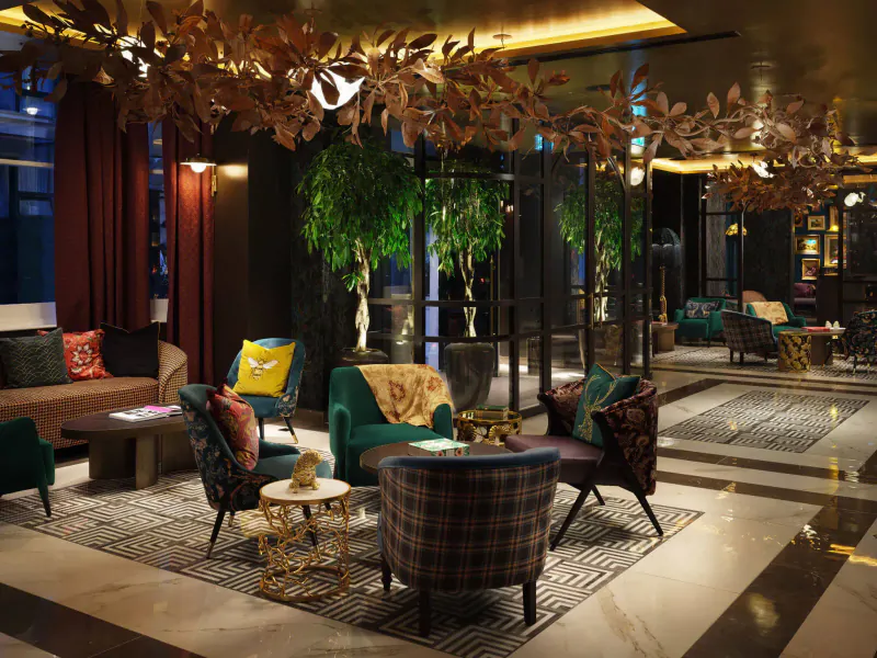 Elegant lobby of The Other House with plush patterned armchairs, gold leaf ceiling sculpture, potted tree, and marble floors.