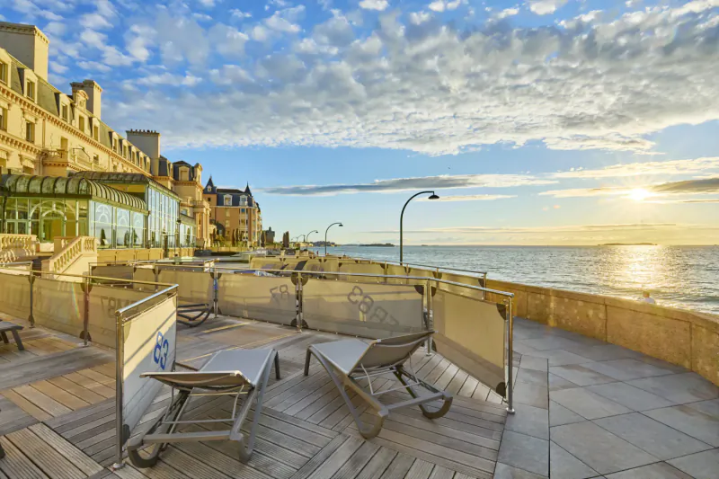 Sunset over Saint-Malo seafront: luxury hotel with lounge chairs on promenade by ocean.