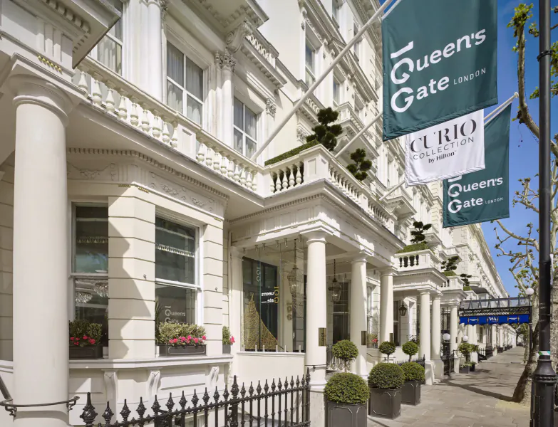 Elegant white facade of 100 Queen's Gate Hotel with green banners, columns, flower boxes, and plants on London street.