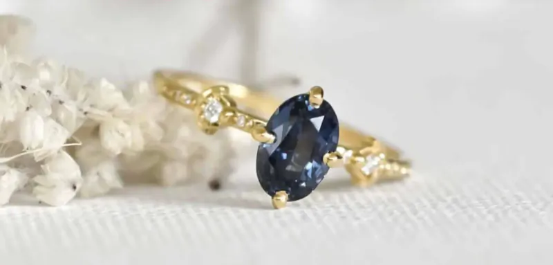 Close-up of gold ring with oval navy blue gemstone and diamond accents on white surface with dried flowers