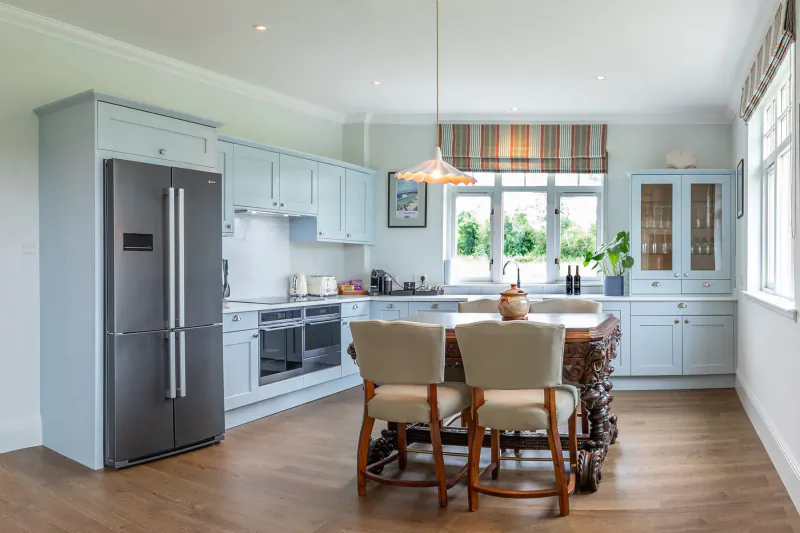 Elegant light blue kitchen in luxury manor home with wooden dining table, chairs, stainless fridge, and garden window view.