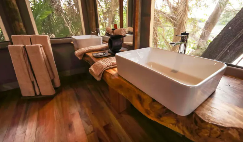 Luxury treehouse bathroom with white vessel sink on live-edge wood vanity, towels, and jungle views through windows.