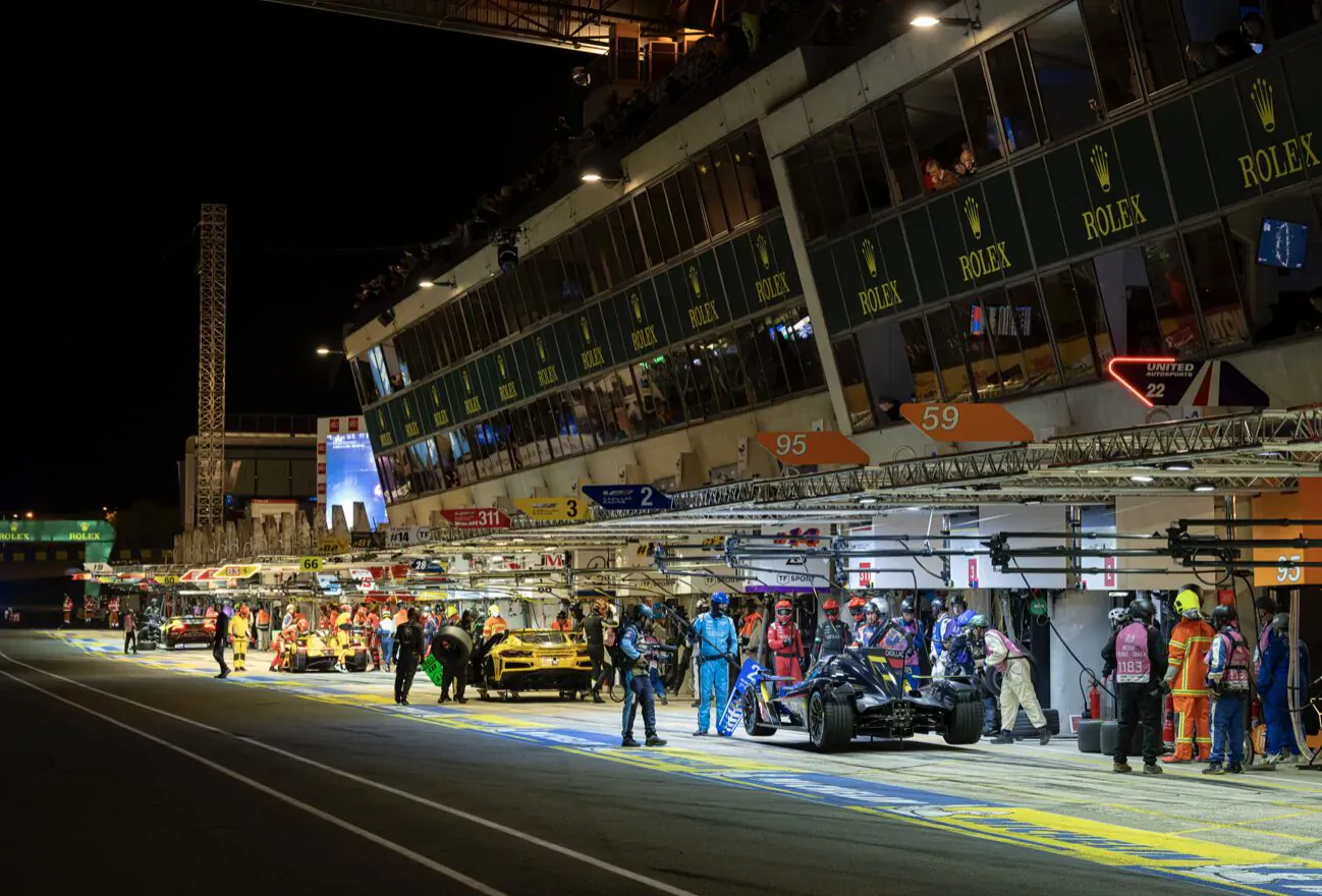 Nighttime 24 Hours of Le Mans pit lane with Rolex-branded garages, race cars, crew in helmets and jumpsuits.