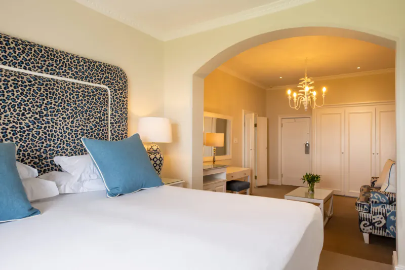 Elegant hotel bedroom at The Marine Hotel Hermanus with blue patterned headboard, white bed, cyan pillows, lamps, and arched doorway to lounge.