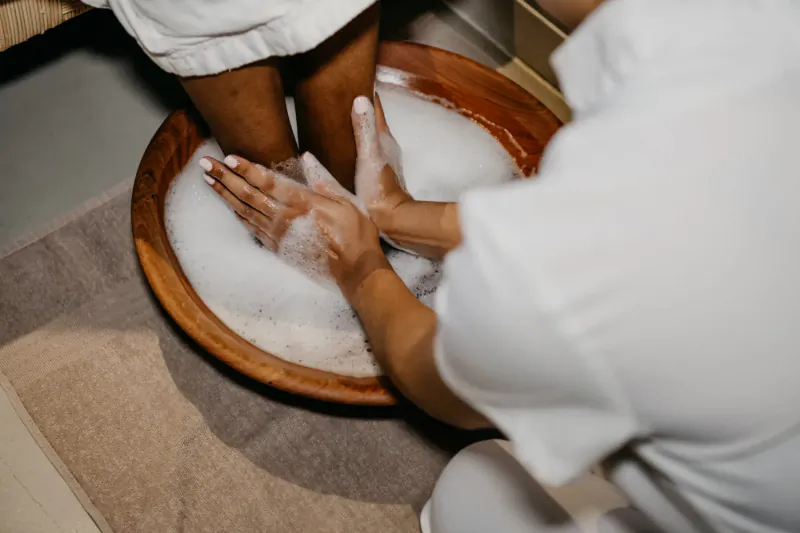 Therapist in white uniform washing client's feet in wooden bowl filled with soapy water at spa.