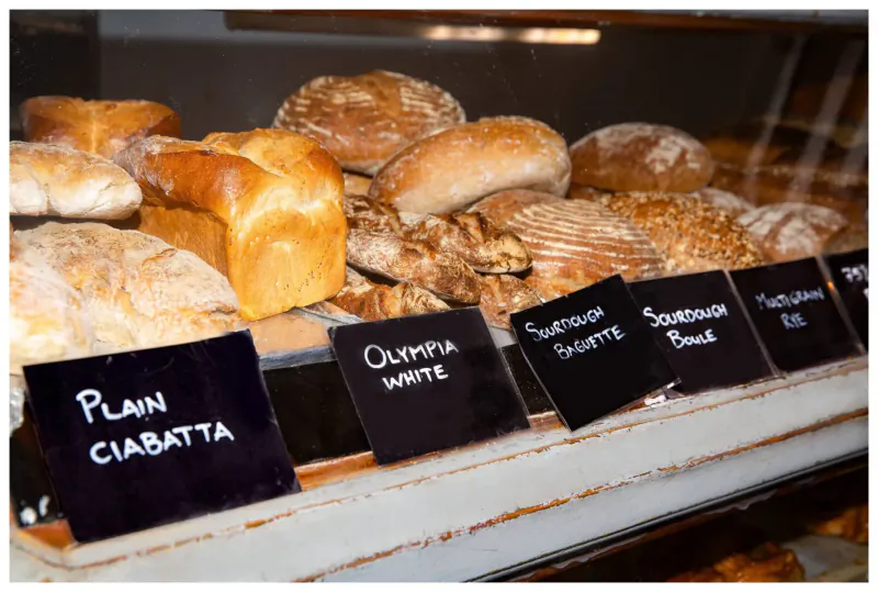 Bakery display case with labeled breads: Plain Ciabatta, Olympia White, Sourdough, Seeded Sourdough, and Rye.