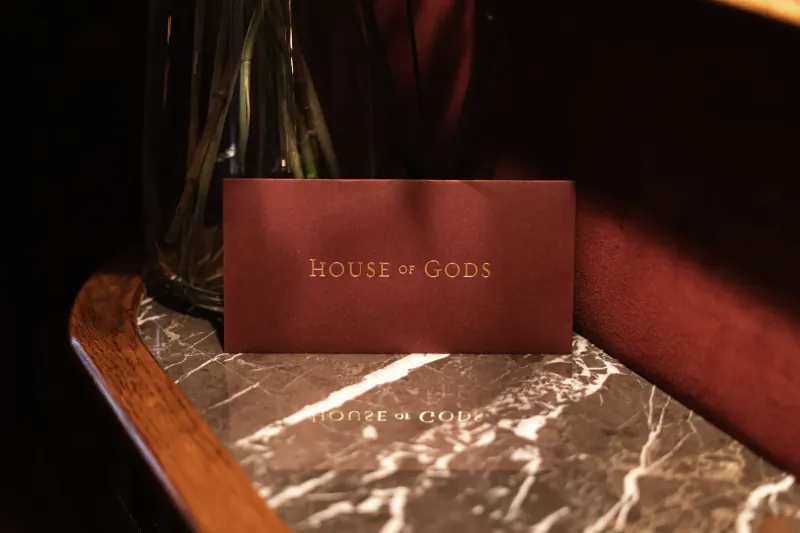 Red 'HOUSE OF GODS' card on marble table beside green vase, in luxurious setting