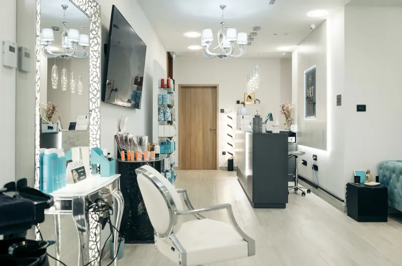 Modern spa salon interior with white stylist chair, vanity mirror, beauty products, TV, and chandelier lighting.