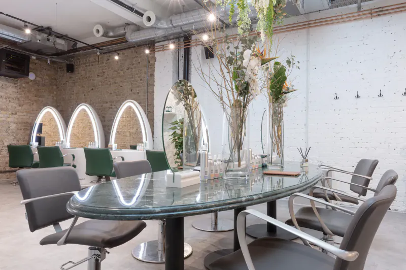 Modern salon interior with arched mirrors, green chairs around glass table with vases, plants and exposed brick walls.