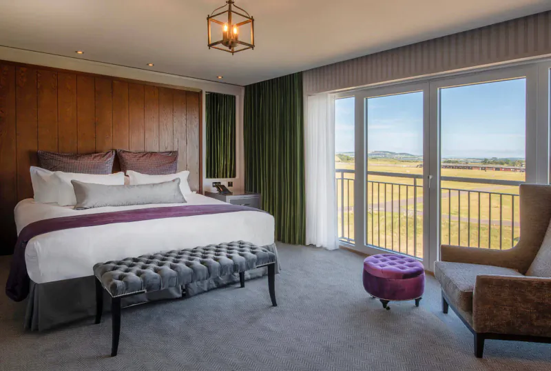 Luxurious hotel bedroom with king bed, balcony view of golf course and fields, wood paneling, and plush seating.