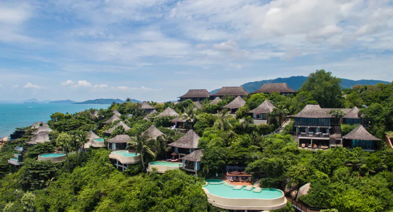 Aerial view of Silavadee Pool Spa Resort: thatched-roof villas nestled in lush greenery on a hillside overlooking the sea and infinity pool.
