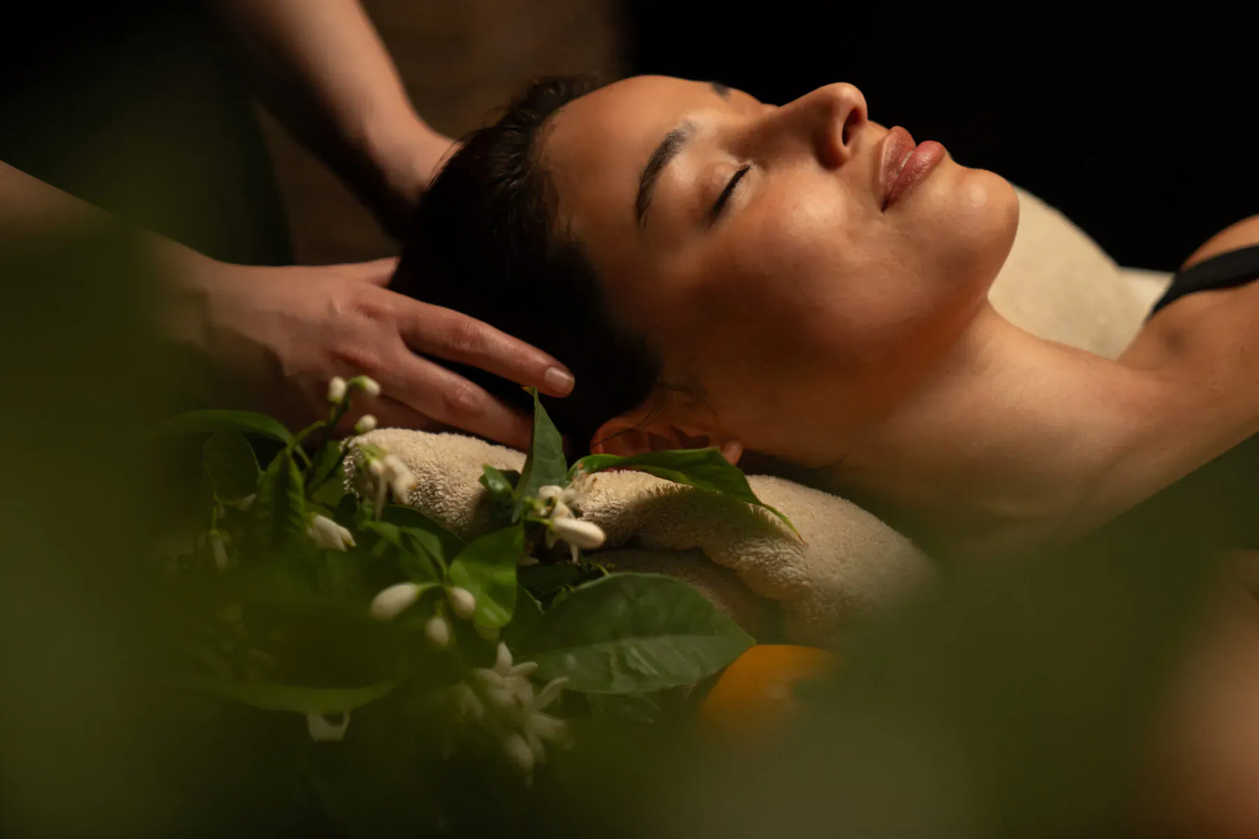 Close-up of woman receiving head massage, eyes closed relaxed, surrounded by orange blossoms and leaves in spa setting