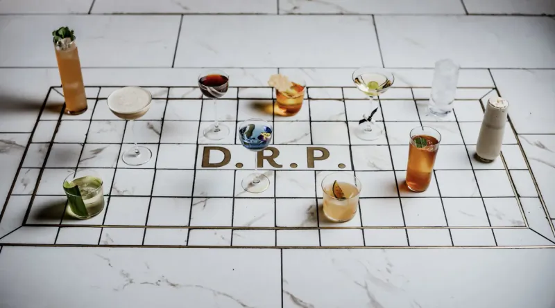 Assortment of colorful cocktails on grid tray over 'D.R.P.' text, with limes and candle on white tile floor
