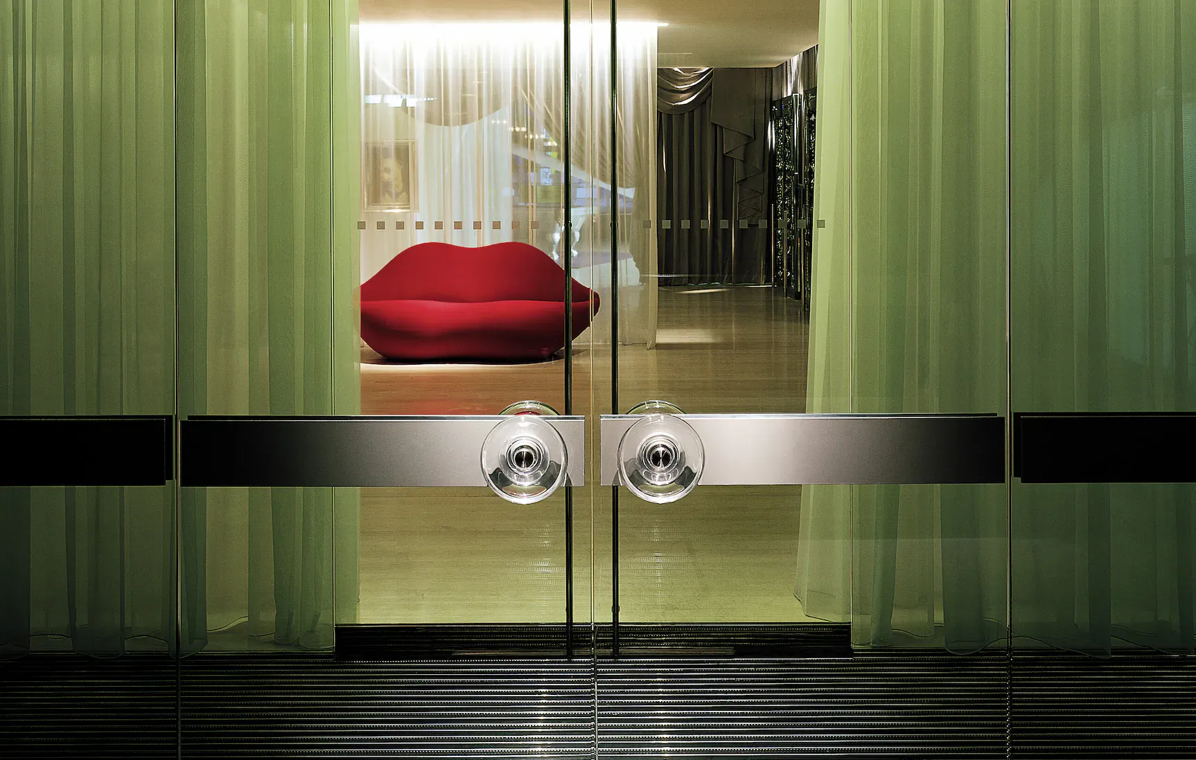 Glass entrance doors with green curtains framing red lips-shaped sofa in Sanderson Hotel lobby