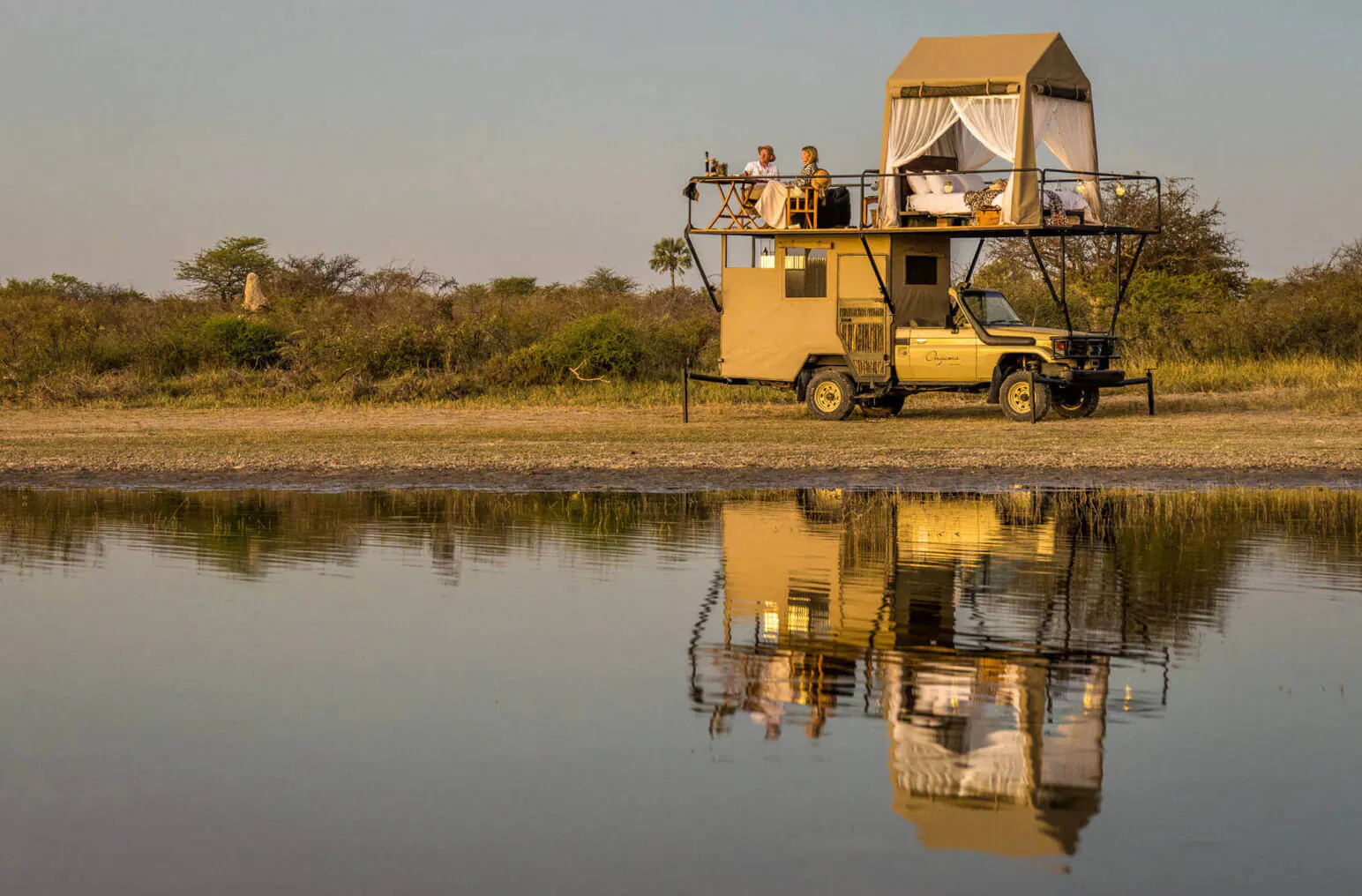 Yellow 4x4 safari vehicle with rooftop tent on platform, people lounging above, reflected in Etosha waterside.