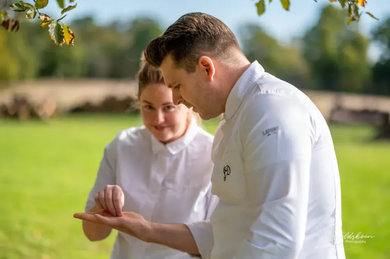 Male chef in white uniform shows small item on palm to smiling female chef outdoors on green lawn amid trees