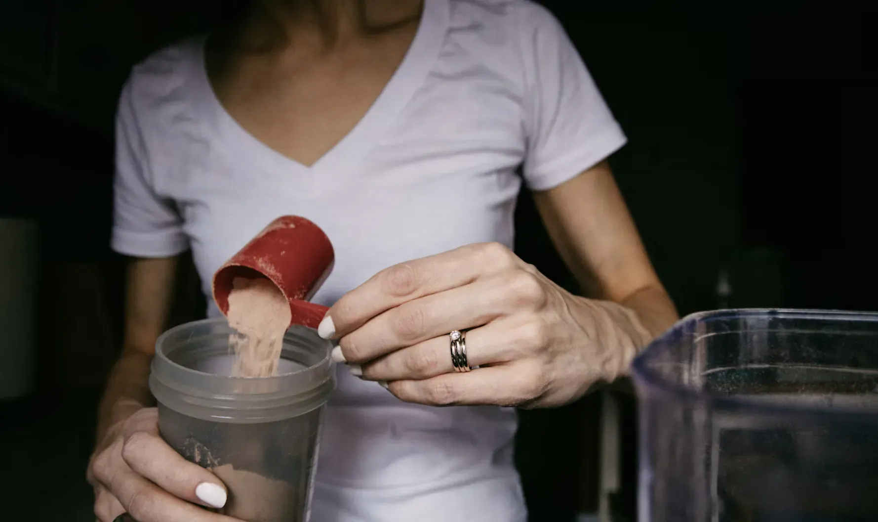 Woman in white V-neck top pouring red scoop of protein powder into clear shaker bottle