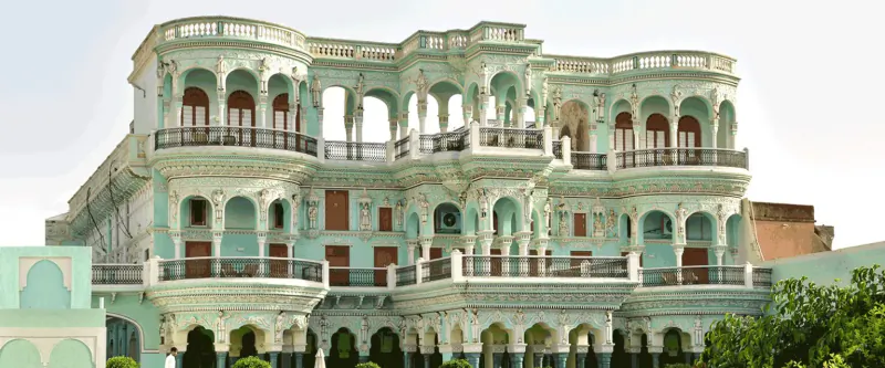 Majestic mint-green multi-story Rajasthani haveli with ornate arches, balconies, and red doors amid greenery.