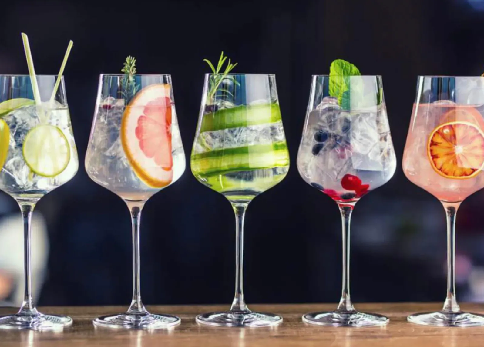 Row of five elegant gin cocktails garnished with lime, grapefruit, cucumber, and berries on a wooden bar.