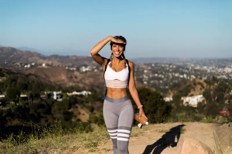 Fitness entrepreneur Lilly Sabri shades her eyes on a sunny LA hilltop trail, wearing white sports bra and striped leggings, holding water bottle.
