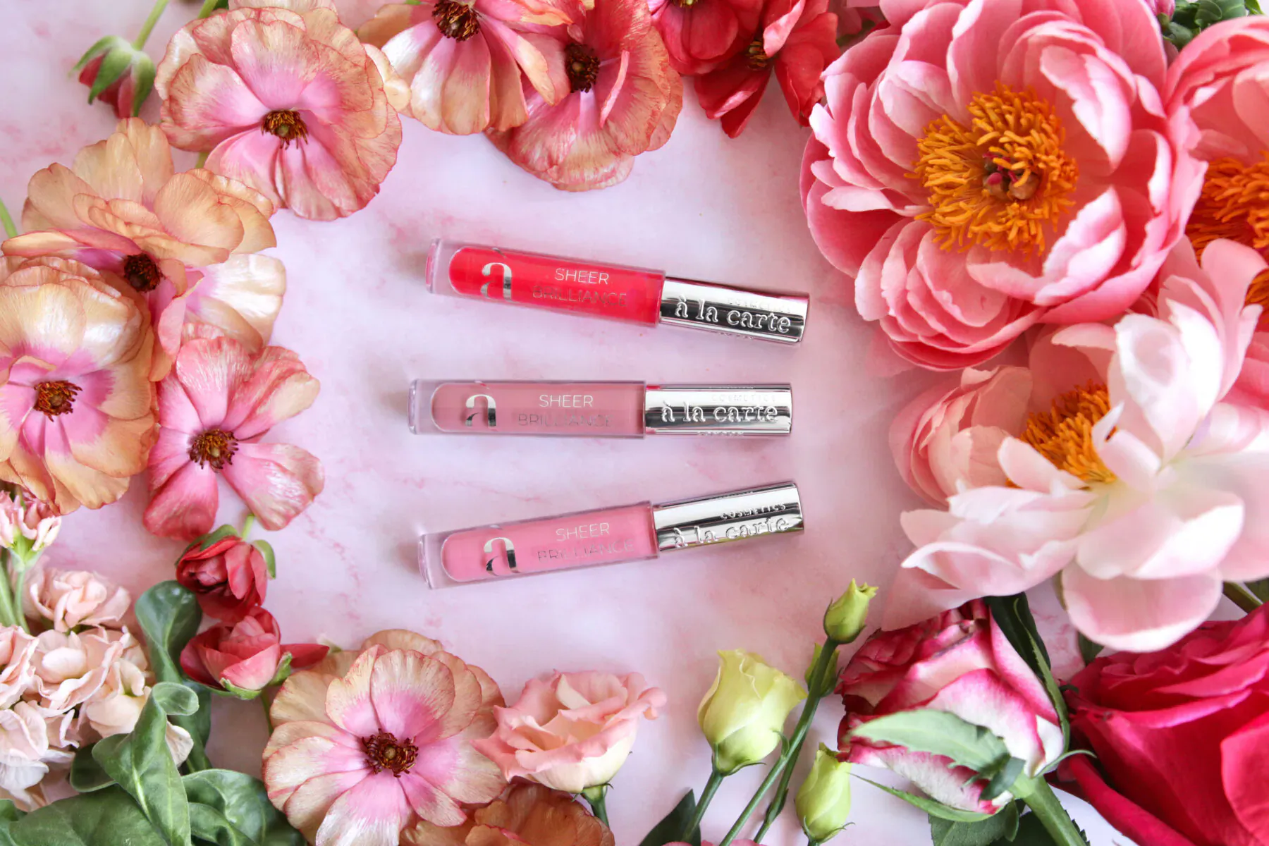 Three Bobbi Brown lip glosses in pink shades arranged amid pink peony flowers for Chelsea in Bloom 2025